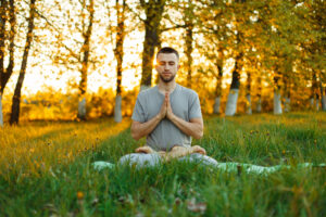 Man meditating in lotus pose with hands in prayer amidst an autumnal forest, embodying tranquility and mindfulness.