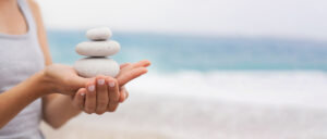 A person holding a stack of three smooth, white stones in their hand against a serene beach backdrop, symbolizing balance and harmony.