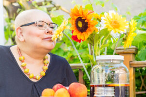 A woman with glasses and a vibrant necklace enjoys the fragrance of colorful flowers in a sunny garden, symbolizing resilience and the essence of womanhood.