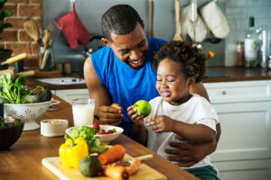 A happy father and his young child enjoying a healthy meal together in a kitchen, surrounded by fresh vegetables and fruits.