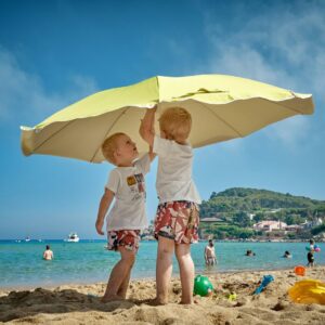 Two young boys playing under a large beach umbrella on a sunny beach, emphasizing sun safety and skin protection for children.