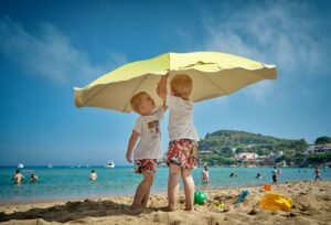Two young boys playing under a large beach umbrella on a sunny beach, emphasizing sun safety and skin protection for children.