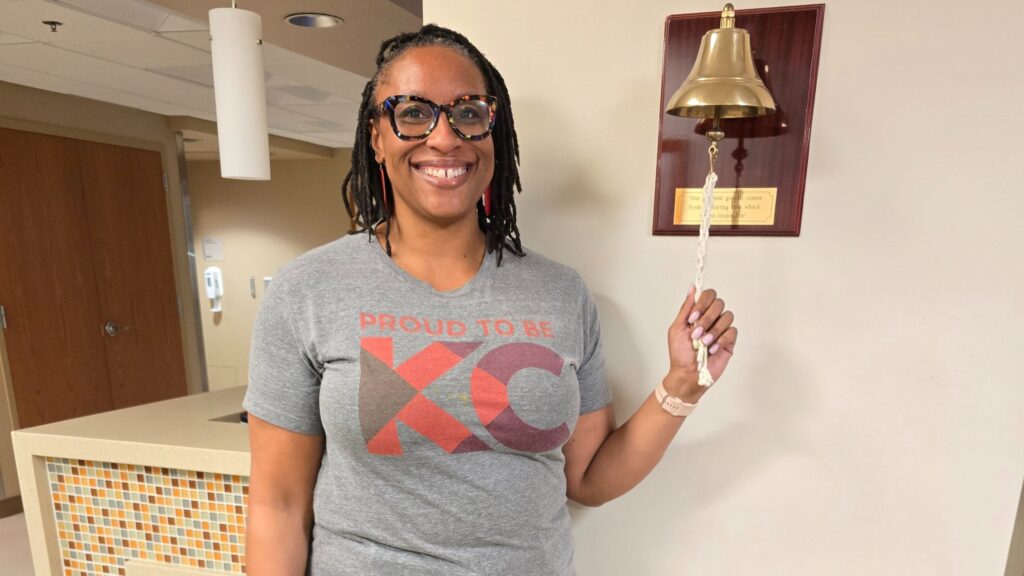 African-American woman smiling and ringing a bell at a hospital after completing cancer treatment, wearing a "Proud to be KC" t-shirt.