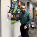 Man in glasses smiling while ringing a celebration bell in a clinic corridor with a "HEAL HEALTHY HABIT" poster on the wall.